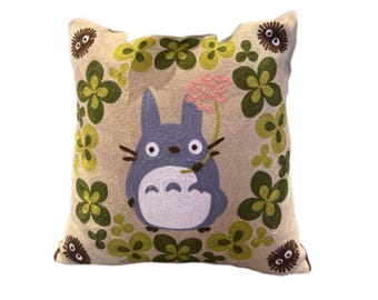 May include: Embroidered square pillow featuring a blue Totoro character holding a pink flower. The pillow has a beige background with green and yellow clover-like designs and small brown, round characters. A decorative accent for a bed or couch.