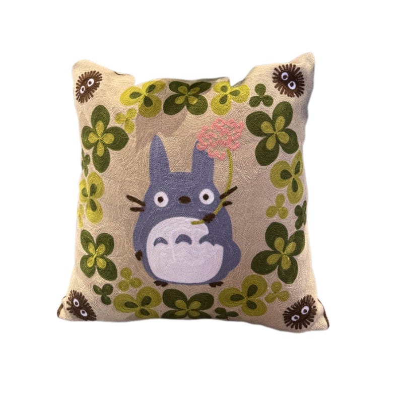 Puede incluir: Coj&iacute;n cuadrado bordado con un personaje Totoro azul sosteniendo una flor rosa. El coj&iacute;n tiene un fondo beige con dise&ntilde;os de tr&eacute;boles verdes y amarillos y peque&ntilde;os personajes redondos marrones. Un acento decorativo para una cama o sof&aacute;.