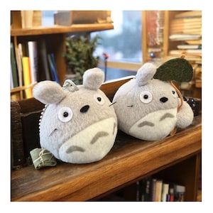K&ouml;nnte beinhalten: Zwei pl&uuml;schgraue Totoro-inspirierte Schl&uuml;sselanh&auml;nger mit wei&szlig;en B&auml;uchen und gro&szlig;en Augen. Ein Schl&uuml;sselanh&auml;nger hat ein gr&uuml;nes Blatt befestigt. Die Schl&uuml;sselanh&auml;nger befinden sich auf einem Holzregal, was auf ein Sammlerst&uuml;ck oder Geschenk hindeutet.