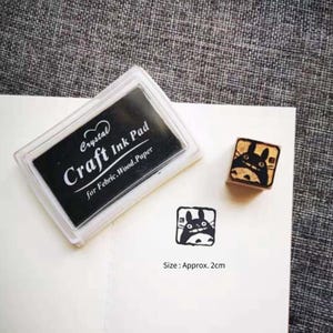 May include: A black ink pad labelled "Crystal Craft Ink Pad for Fabric, Wood, Paper" with a clear plastic case. Next to it is a small wooden stamp with a black and white illustration of a cartoon character with large ears.