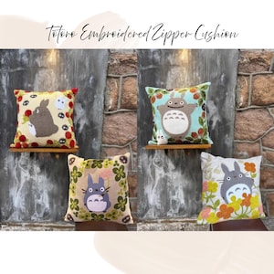 Puede incluir: Cuatro cojines bordados con cremallera con dise&ntilde;os de Totoro. Cada coj&iacute;n cuadrado muestra un personaje de Totoro diferente con fondos florales e inspirados en la naturaleza. Los cojines se exhiben en estantes de madera contra una pared de piedra.
