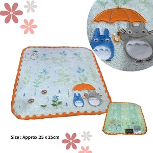 May include: A light blue, approximately 9.8 x 9.8 inch, square towel with an orange scalloped border. The towel features embroidered images of Totoro characters under an orange umbrella, with floral and rain drop details.