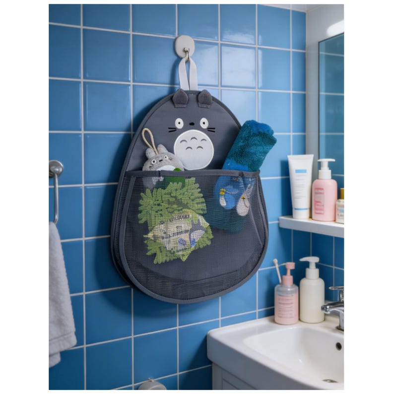Puede incluir: Un organizador colgante gris con tem&aacute;tica de personaje con un bolsillo de malla, colgado en una pared de azulejos azules. El organizador tiene la forma de un personaje de dibujos animados y contiene varios art&iacute;culos de ba&ntilde;o, incluyendo toallitas y un peluche.