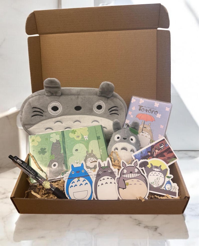 May include: A brown cardboard box filled with My Neighbor Totoro themed items. The box contains a gray plush Totoro eye mask, two green notebooks with Totoro designs, a gray Totoro keychain, a blue Totoro sticker, a gray Totoro sticker with a flower, a gray Totoro sticker with a toothy grin, a sticker of a scene from the movie, and a black pen.