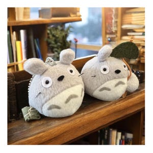 Puede incluir: Dos llaveros de peluche grises inspirados en Totoro. Cada llavero presenta un cuerpo redondo, orejas peque&ntilde;as y una barriga blanca. Uno tiene una hoja verde adjunta. Los llaveros se exhiben en una estanter&iacute;a de madera.