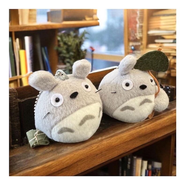 Puede incluir: Dos llaveros de peluche grises inspirados en Totoro. Cada llavero presenta un cuerpo redondo, orejas peque&ntilde;as y una barriga blanca. Uno tiene una hoja verde adjunta. Los llaveros se exhiben en una estanter&iacute;a de madera.