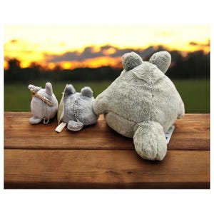 May include: Three gray plush Totoro figures of varying sizes are arranged on a wooden surface. The largest plush has a round body and a small tail. The smaller figures have keychains attached. The background features a sunset.