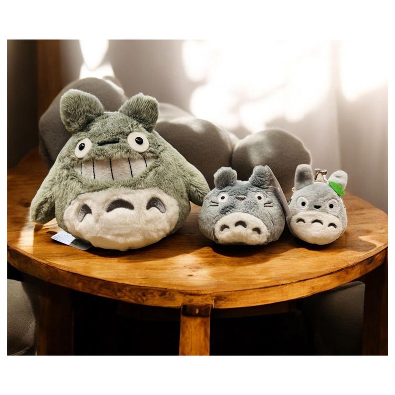 May include: A collection of plush Totoro characters in various sizes. The largest is a light green color with a smiling face and white belly. Two smaller gray Totoro plushies are also present, one with a green leaf.