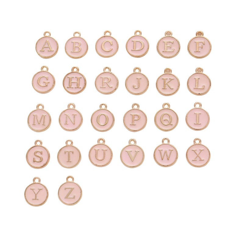 May include: A collection of gold-colored alphabet charms, each with a light pink enamel background. The charms are round with a loop at the top for attaching to jewelry. The letters A through Z are displayed.