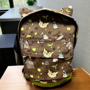 May include: A brown backpack featuring a repeating pattern of cartoon characters, leaves, and insects. The backpack has a front pocket, green trim, and cat-like ears. It is on a wooden surface.