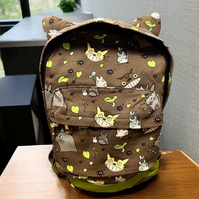 May include: A brown backpack featuring a repeating pattern of cartoon characters, leaves, and insects. The backpack has a front pocket, green trim, and cat-like ears. It is on a wooden surface.