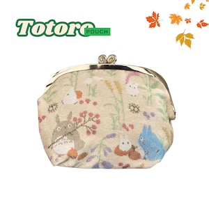 May include: A beige pouch with a metal clasp, featuring a whimsical pattern of cartoon characters, flowers, and foliage. The text "Totoro Pouch" is displayed in green and white. The pouch has a gold-colored metal frame.