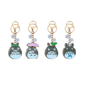 May include: Four keychains featuring a cartoon character with gray bodies and light blue bellies. Each keychain has a gold-colored clasp and a charm with the letter "M". One character holds a pink umbrella, another has a green leaf.