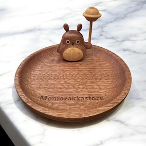 Op de afbeelding: Een houten serveerschaal met een gesneden Totoro-figuur en een accessoire in de vorm van een lolly. De bruine schaal heeft de tekst "Momozakkastore" gegraveerd. De Totoro-figuur is bruin en beige.