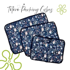 Puede incluir: Tres cubos de embalaje azul marino con un patr&oacute;n caprichoso con personajes Totoro blancos y elementos florales. Los cubos son de diferentes tama&ntilde;os, con un borde azul oscuro y el texto "Totoro Packing Cubes".