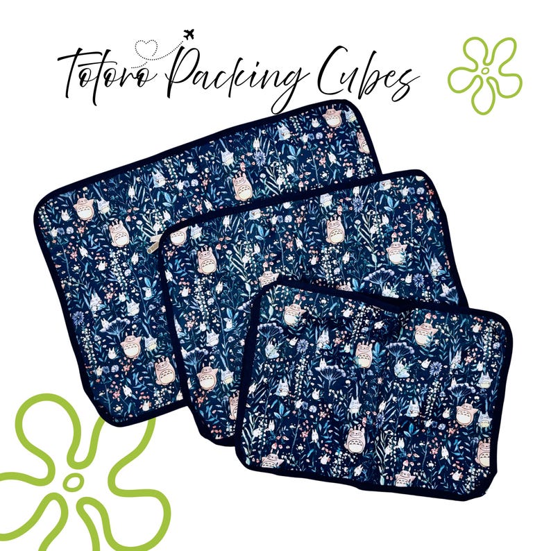 Puede incluir: Tres cubos de embalaje azul marino con un patr&oacute;n caprichoso con personajes Totoro blancos y elementos florales. Los cubos son de diferentes tama&ntilde;os, con un borde azul oscuro y el texto "Totoro Packing Cubes".