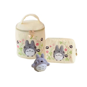 May include: A set of three items: a small plush toy and two cream-colored corduroy cosmetic bags. The bags and toy feature embroidered images of a gray cartoon character with floral accents. The bags have gold zippers.