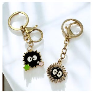 May include: Two gold keychains featuring black and white cartoon characters. The first keychain has a black character with large eyes and green and pink stars. The second keychain has a black character with large eyes and a gold outline.