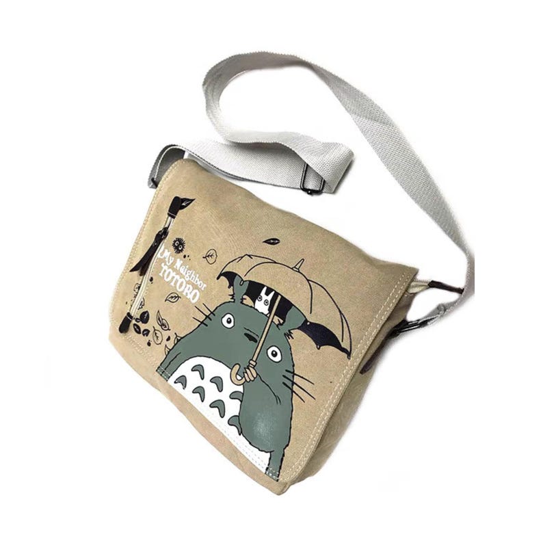 May include: Beige canvas messenger bag featuring a graphic of Totoro characters under an umbrella. The bag has a white adjustable strap and the text "My Neighbor Totoro". The bag is designed with a flap closure.