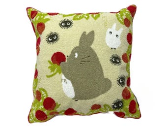 May include: A cream-colored throw pillow with a plush texture features a cartoon character with large ears and a white bunny. The pillow is decorated with red berries, green leaves, and black and white cartoon creatures.