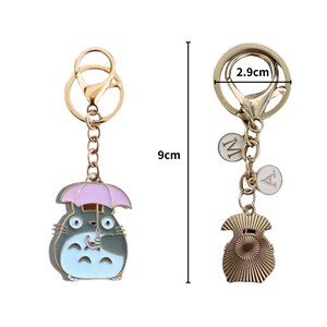 May include: A gold-coloured keyring featuring a cartoon character holding an umbrella. The keyring is 9 cm long and has a 2.9 cm wide clasp. The character is grey, blue, and white with a pink umbrella. Includes two small charms.