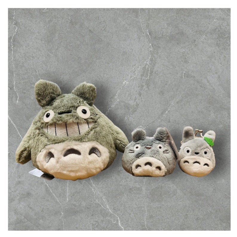 May include: Three plush Totoro figures in varying sizes. The largest is olive green with a cream belly and a wide grin. The smaller ones are gray, with a keychain attached to the smallest. All have large eyes and a soft texture.