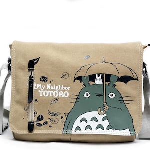 May include: Beige canvas messenger bag featuring a graphic of Totoro from "My Neighbor Totoro" holding an umbrella. The bag has a gray adjustable strap, a front flap, and a zipper pocket. The text "My Neighbor Totoro" is printed on the front.
