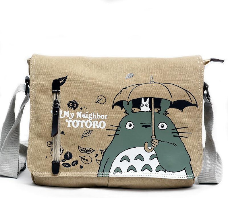 May include: Beige canvas messenger bag featuring a graphic of Totoro from "My Neighbor Totoro" holding an umbrella. The bag has a gray adjustable strap, a front flap, and a zipper pocket. The text "My Neighbor Totoro" is printed on the front.