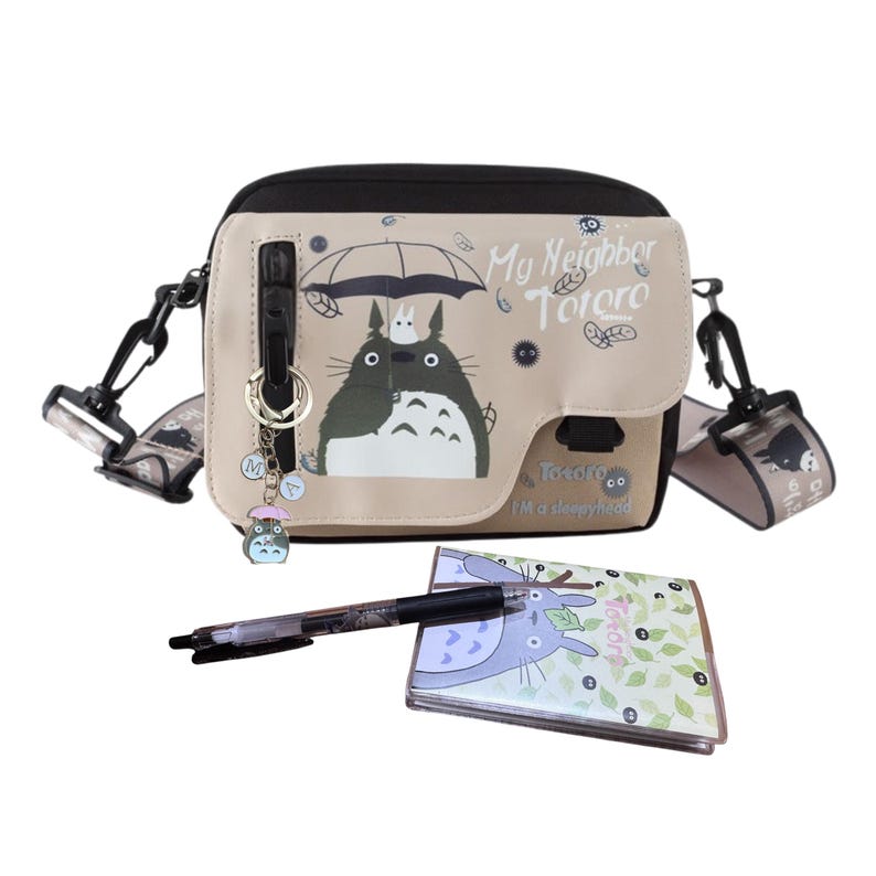 May include: A beige and black crossbody bag with a Totoro graphic, a matching notebook, and a pen. The bag has an adjustable strap and a charm. The text "My Neighbor Totoro" is printed on the bag. The notebook and pen are on a white surface.