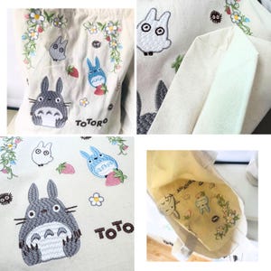 May include: A white canvas tote bag with embroidered images of Totoro, a small white bunny, and a blue cat-like creature. The bag has the word "TOTORO" embroidered on the front. The inside of the bag is lined with a yellow fabric with a similar embroidery design.