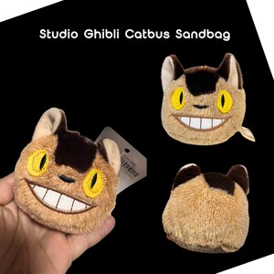 May include: Studio Ghibli Catbus sandbag. Plush sandbag shaped like the Catbus from the animated film. The sandbag is tan with brown and yellow accents, featuring a smiling face with large yellow eyes and a wide grin.