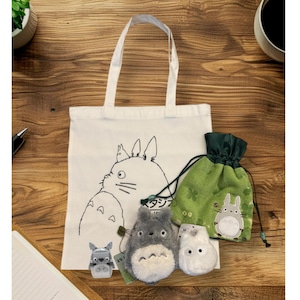 May include: A white tote bag featuring a black outline illustration of a cartoon character, with plush toys and a green drawstring bag. The plush toys include a gray Totoro, a white Soot Sprite, and a gray robot-like figure.