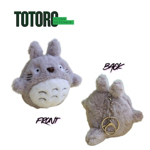 May include: A plush Totoro keychain in light gray with a white belly and a brown nose. The front view shows the face, while the back view reveals a keyring. The text "TOTORO PLUSH KEYCHAIN" is at the top.
