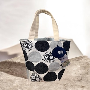 May include: A small tote bag with a repeating pattern of black, gray, and white circular shapes with stylized eye designs. The bag has cream-colored handles and a small, dark blue plush charm with googly eyes attached.