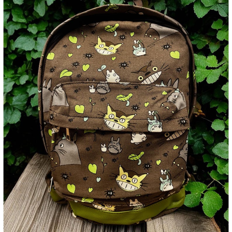 May include: Brown backpack with a repeating pattern of cartoon characters, including a large cat with yellow eyes and a wide grin. The backpack has a front pocket and green accents on the bottom and straps. The background is a natural green.