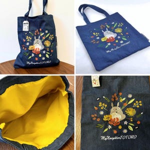May include: A denim tote bag featuring an embroidered design of a cartoon character with floral accents. The bag has a yellow interior and the text "My Neighbor Totoro". The bag has two handles for carrying.