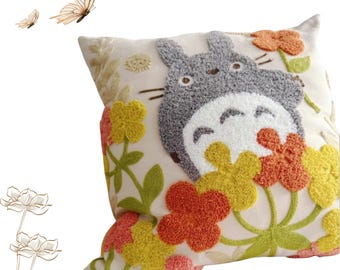 May include: Embroidered pillow featuring a gray Totoro character with a white belly, surrounded by colorful flowers in shades of orange, yellow, and pink. The pillow has a cream-colored background and green embroidered leaves.
