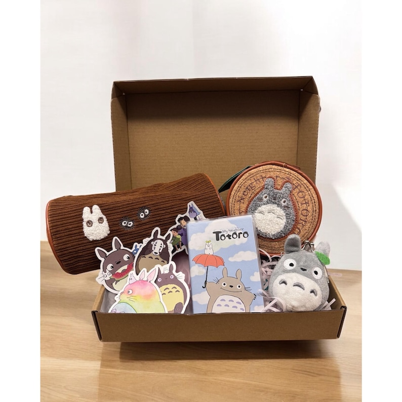 K&ouml;nnte beinhalten: Eine braune Pappbox gef&uuml;llt mit Artikeln zum Thema Mein Nachbar Totoro. Die Box enth&auml;lt eine braune Pl&uuml;sch-Federmappe, eine runde Geldb&ouml;rse mit Totoros Gesicht, einen kleinen Pl&uuml;sch-Totoro-Schl&uuml;sselanh&auml;nger, eine Sticker-Tafel mit Totoro und anderen Figuren und ein kleines Notizbuch mit dem Text "Mein Nachbar Totoro" auf dem Cover.