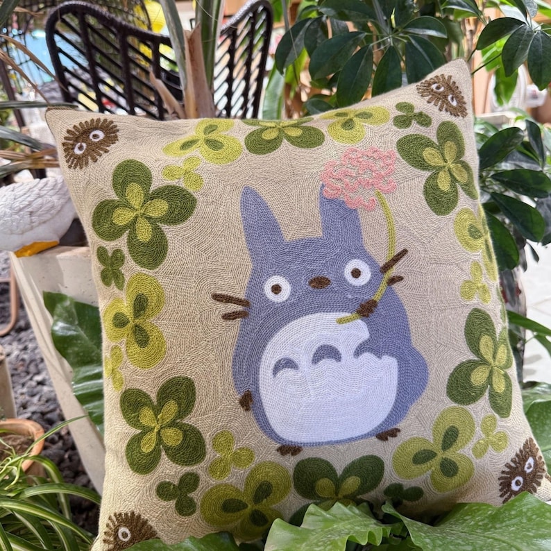 Puede incluir: Coj&iacute;n cuadrado beige con un dise&ntilde;o bordado que presenta un personaje Totoro azul sosteniendo una flor rosa. El coj&iacute;n est&aacute; decorado con formas de tr&eacute;bol verdes y amarillas y acentos circulares marrones.