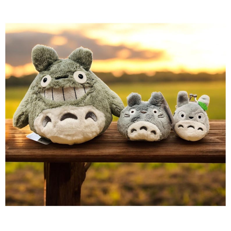 May include: Three plush Totoro figures in varying sizes sit on a wooden surface. The largest is a light green color with a wide grin and white belly. The smaller ones are gray, one with a keychain. The background is a blurred sunset.
