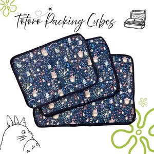Puede incluir: Tres cubos de embalaje azul marino con un estampado floral y del personaje Totoro. Los cubos tienen un borde negro y est&aacute;n apilados. El texto "Totoro Packing Cubes" est&aacute; escrito en cursiva en la parte superior de la imagen.