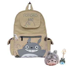 Könnte beinhalten: Beigefarbener Rucksack mit einem großen grauen Totoro-Motiv und dem Text "MY NEIGHBOUR TOTORO". Enthält einen Totoro-Plüsch und einen Totoro-Schlüsselanhänger. Der Rucksack hat Seitentaschen und eine Reißverschlusstasche vorne.