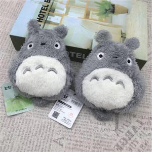 May include: Two gray plush Totoro plushies with white bellies and black eyes. The plushies are sitting on a patterned surface.