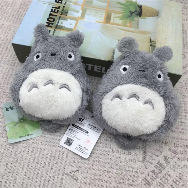 May include: Two gray plush Totoro plushies with white bellies and black eyes. The plushies are sitting on a patterned surface.