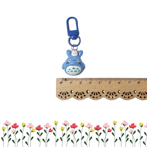 May include: A blue Totoro keyring with a blue clasp and ring. The keyring is approximately 4 cm tall. The Totoro figure has a white belly with a pattern of triangles and a small white figure on top.