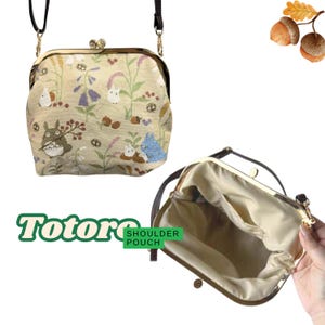 May include: A beige shoulder pouch with a clasp closure, featuring a floral and Totoro character design. The bag has a brown strap and gold-colored hardware. The text "Totoro Shoulder Pouch" is displayed in green and white.