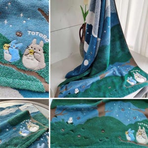 May include: A blue and green plush towel featuring an embroidered design of Totoro characters in a forest scene. The towel has a night sky design with stars and a moon. The word "TOTORO" is embroidered on the towel.
