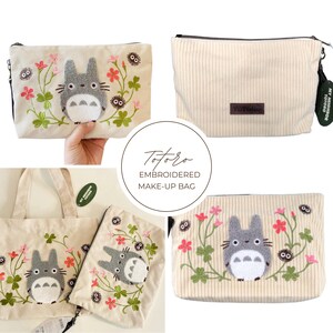 May include: Cream corduroy makeup bag with an embroidered Totoro design. The design features a grey Totoro character surrounded by pink flowers and green vines. The bag has a black zip and a small tag.