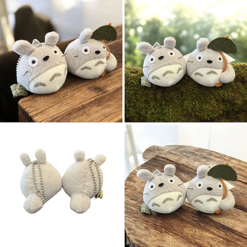 Puede incluir: Dos llaveros de peluche de Totoro. Los peluches grises y blancos tienen cuerpos redondos, orejas peque&ntilde;as y ojos negros. Uno tiene un accesorio de hoja verde. Se exhiben sobre una superficie de madera.