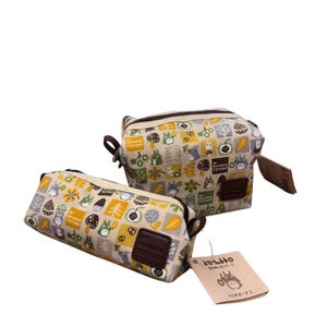 Op de afbeelding: Twee beige My Neighbor Totoro-etuis met een herhalend patroon van Totoro-personages, bloemen en bladeren in geel, grijs en groen. De etuis hebben bruine ritsen, handvatten en leren patches. Eén etui is rechthoekig, de andere is vierkant.